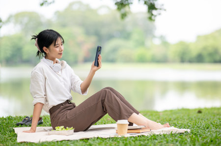 Asian woman in shirt looking at phone or taking a photo as sitting leaning back on picnic mat laying on grass. Relaxing Outdoor, Chilling in the park.の写真素材