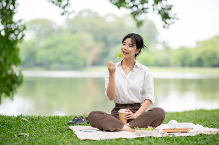 Asian woman in white shirt smiling happily as holding fork eating salad while sitting on picnic mat...