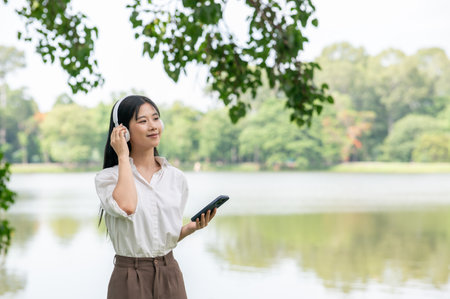 Asian woman in white shirt listening to music in headphones while standing or walking around the lake or river. Relaxing Outdoor, Chilling in park.の写真素材