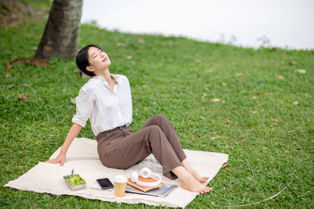 Asian woman in white shirt deep breathing in nature while sitting leaning back on picnic mat laying on grass. Relaxing Outdoor, Chilling in the park.の写真素材