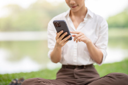 Close up of a woman in white shirt holding and looking at her smartphone with blurred grass and water background. Relaxing Outdoor, Chilling in park.の写真素材