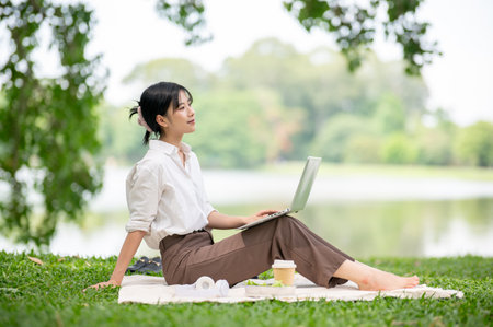Asian woman in white shirt looking away from her laptop while sitting knee up on picnic mat laying on grass. Relaxing Outdoor, Chilling in park.の写真素材
