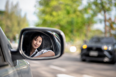 Side mirror shot of smiling asian woman in white shirt in driver seat of a car parked at the lot or roadside. Picking up a friend, Waiting for family.の写真素材