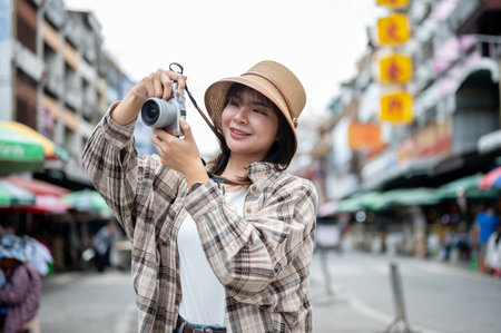 Asian tourist woman wearing hat holding camera with both hands taking photos while standing by the road of local market. Walking street, Old town trip.の写真素材