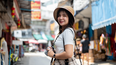 Asian woman wearing hat smiling while holding her camera with both hands standing in the local old...