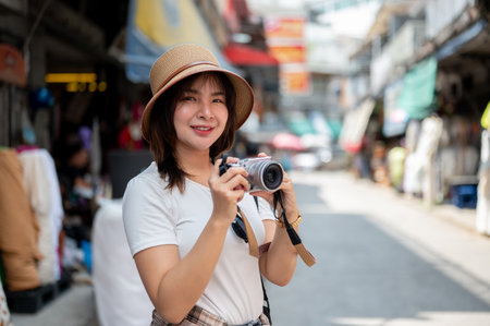 Asian tourist woman wearing hat smiling while holding a camera taking photos around the old market. Tourist spot, Walking street, Old town trip.の写真素材