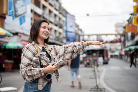 Asian tourist woman holding phone and raising one arm to catch a taxi while standing by the road in...