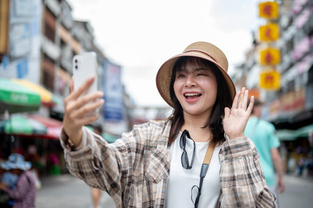 Asian tourist woman with hat holding phone waving a hand during video call while standing by the...