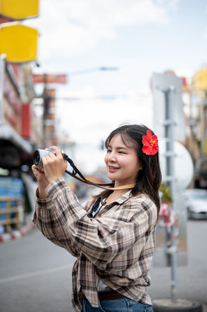 Smiling asian tourist woman with red hibiscus flower on hair taking photo on camera along the road in local market. Walking street, Old town trip.の写真素材