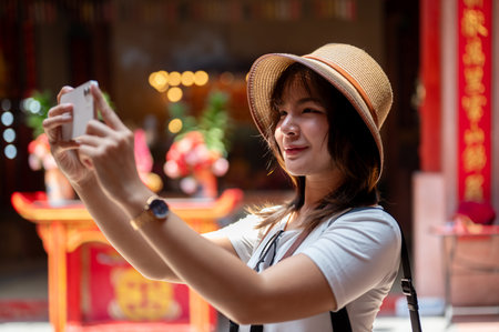 Asian woman wearing hat holding smartphone with both hands taking photos in the Chinese shrine...