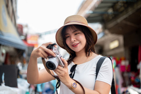 Asian tourist woman wearing hat smiling while holding camera checking photos taken in the local market. Tourist spot, Walking street, Old town trip.の写真素材