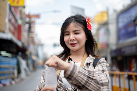 Asian tourist woman with red hibiscus flower on hair opening a cool water bottle to drink as standing in local market. Walking street, Old town trip.の写真素材