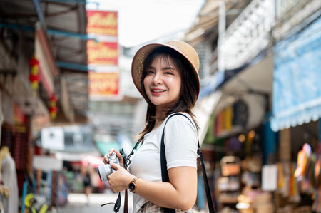 Asian tourist woman wearing hat smiling while holding camera with both hands standing in the local market. Tourist spot, Walking street, Old town trip.の写真素材