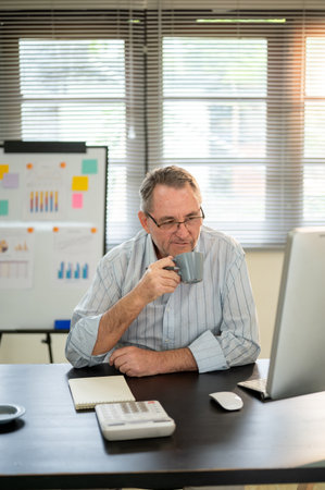 Old white man wearing glasses holding coffee cup and looking at computer on working table in office.の写真素材