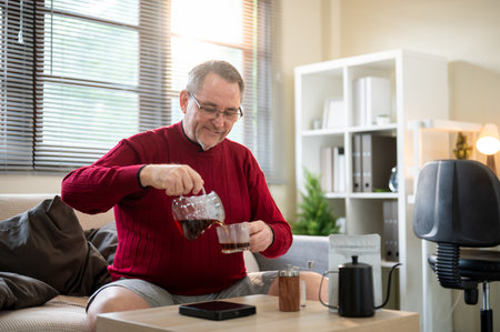 Old white caucasian man wearing glasses pouring freshly brew coffee in the dripper into a cup on sofa in a living room. Pour over or Hand Drip.の写真素材