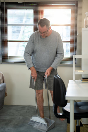 Old white caucasian man wearing glasses standing holding a broom sweeping the dust on a carpet and floor in his room. Housework, household chores.の写真素材