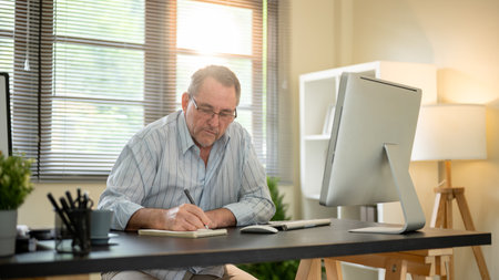 Old white caucasian man wearings eyeglasses writing in notebook with a pen on working table in his office. Company job, Boss or Senior manager.の写真素材