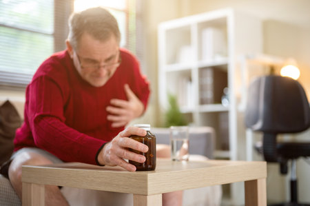 Old white man getting heart attack or cardiac arrest as grabbing medicine bottle on table in living room. Health problems, Disease, Illness.の写真素材