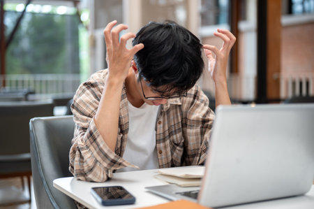 Man in plaid shirt raising curl up hands thinking or stressing in front of laptop and notebook sitting at cafe table. Study or Working, Going Outside.の写真素材