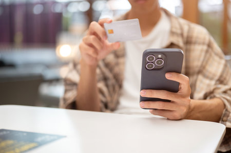 Close up of man holding smartphone and looking closely at credit card sitting at white table in cafe.の写真素材