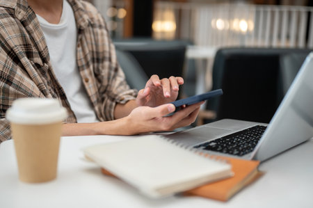 Close up of a man hand holding and touching on phone over laptop with coffee and notebook aside sitting at cafe table. Study or Working, Going Outside.の写真素材