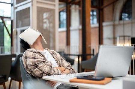 Man in plaid shirt sitting leaning against chair with book covered his face in front of a laptop at cafe table. Study or Working. Sleeping or Resting.の写真素材