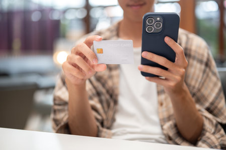 Close up of man holding smartphone and looking at credit card while sitting at white table in cafe.の写真素材
