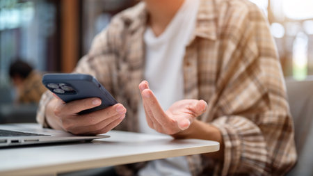 Close up of a man hands holding smartphone over laptop and talking or explaining while sitting at white table in cafe. Study or Working, Going Outside.の写真素材