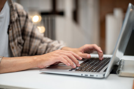 Close up of a man in plaid shirt typing on laptop with both hands while sitting at white table in the cafe. Study or Working, Going Outside.の写真素材