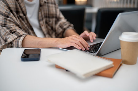 Close up of a man's hands typing and tapping on laptop with phone and notebook while sitting at white table in cafe. Study or Working, Going Outside.の写真素材