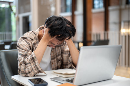 A man in plaid shirt holding grabbing his hair thinking or stressing in front of a laptop and notebook sitting at cafe table. Study or Working Outside.の写真素材