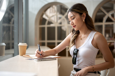 Woman in tank top holding a pen writing in notebook with phone and coffee cup aside sitting at wooden table. Chilling Outside, Working or study.の写真素材