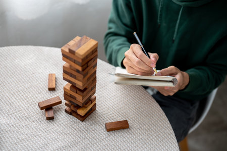 Close up a man holding pen writing in notebook aside a wooden blocks of tower building game on...