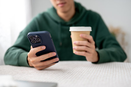 Close up of a man holding a cup of coffee and looking at phone while sitting at table in a cafe....