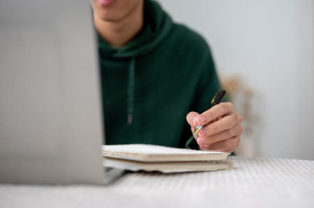 Close up of a man holding pen over notebook while looking at laptop on table in a cafe or library. Working or Study, Doing Assignment, Urban Life.の写真素材