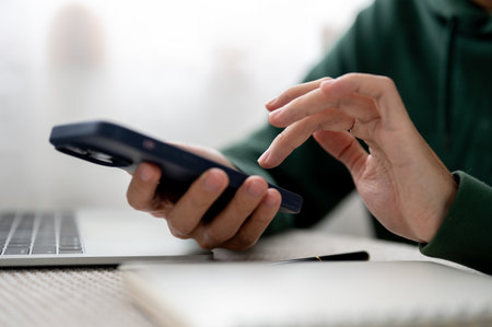 Close up man holding phone and one hand typing aside laptop and notebook at table in cafe or library. Smartphone users, Modern Tech, Urban City Life.の写真素材