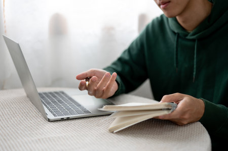 Close up of a man holding pen and notebook while looking at laptop on round table in cafe or library. Working or Study, Doing Assignment, Urban Life.の写真素材