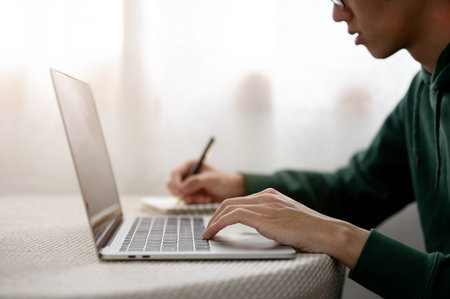 Close up of a man holding pen writing in notebook and typing on laptop as sitting at table in a cafe. Working or Study, Doing Assignment, Urban Life.の写真素材