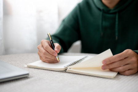 Close up of a man holding pen writing and opening a notebook aside laptop sitting at table in a cafe. Working or Study, Doing Assignment, Urban Life.の写真素材