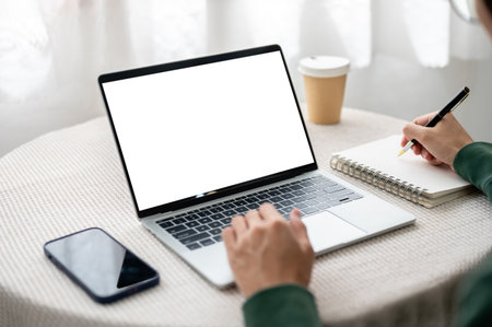 Hand typing on blank screen laptop as writing in notebook with phone on table in cafe or living room. Working or Study, Advertising Mockup, Urban Life.の写真素材