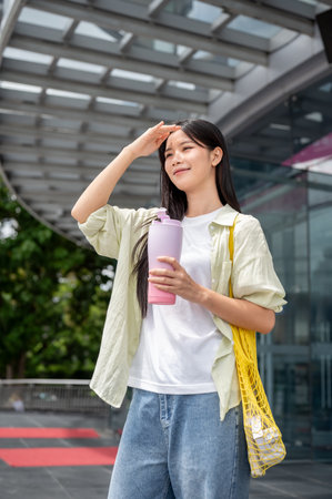 Asian woman with hand blocking sun holding thermos bottle while standing in front of a shopping mall. Casual Clothes, Eco foldable bag, Urban life.の写真素材