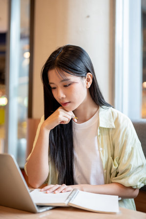 Asian woman student resting head on hand holding notebook looking at laptop on wooden table in cafe. Freelancer, Working or Study, Modern Urban Life.の写真素材