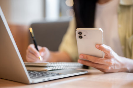 Close up woman student holding pen writing in notebook looking at phone aside laptop on table in cafe. Freelancer, Working or Study, Modern Urban Life.の写真素材