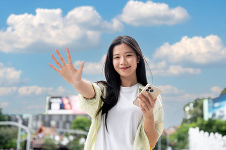 Smiling asian woman holding smartphone and waving a hand while standing outdoor under the blue sky. Casual Clothes, Clear weather, Urban City life.の写真素材