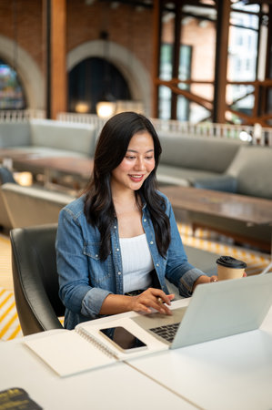 Asian woman student holding coffee typing with pen in hand in front of laptop and book on cafe...