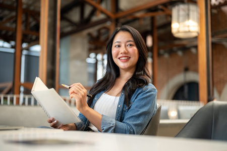 Smiling asian woman holding pen over notebook and looking away at table in a cafe or coffee shop. Working or Study, Freelancer, Urban City Life.の写真素材