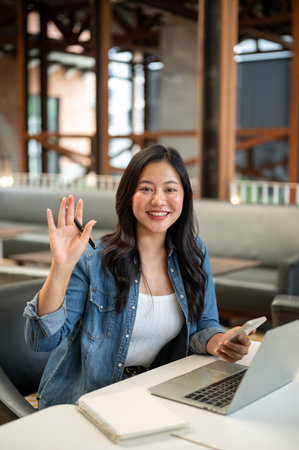 Asian woman holding phone and waving hand with pen in front of laptop and notebook on table in cafe. Online Freelancer, Modern Tech, Urban City Life.の写真素材