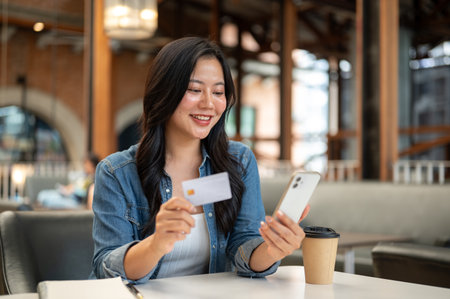 Pretty woman holding credit card and looking at phone aside notebook and coffee at cafe table. Urban Life.の写真素材