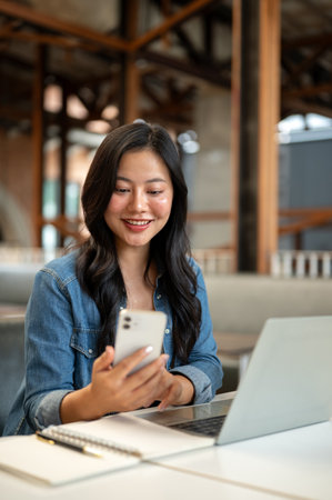 Asian woman student holding and looking at phone over notebook aside laptop sitting at table in...