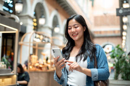 Asian woman holding and looking at phone while walking or standing in shopping mall or indoor market. Travel Alone, Tourist Attractions, Memorable Trip.の写真素材